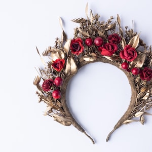 May include: A gold-colored floral crown featuring red roses and berries. The crown is adorned with gold-painted leaves and other botanical elements. The base of the crown is a textured, brown band.