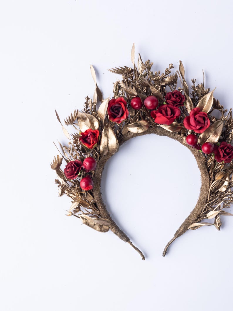 May include: A gold-colored floral crown featuring red roses and berries. The crown is adorned with gold-painted leaves and other botanical elements. The base of the crown is a textured, brown band.