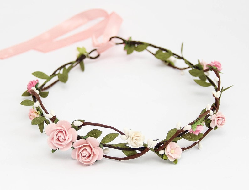 May include: A pink floral crown with small pink roses and white flowers on a brown vine base. The crown is tied with a pink ribbon.