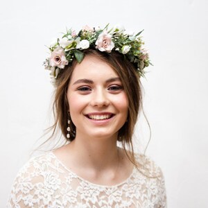 May include: A woman wearing a white lace dress with a floral crown on her head. She is smiling and looking at the camera.