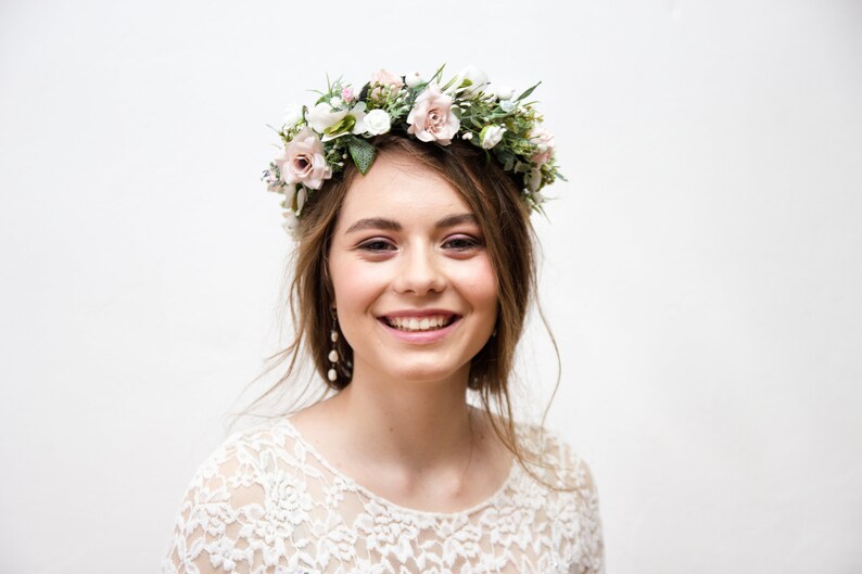 May include: A woman wearing a white lace dress with a floral crown on her head. She is smiling and looking at the camera.