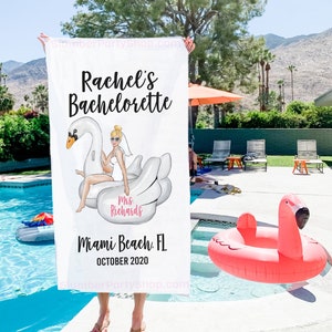 May include: White beach towel with a cartoon illustration of a bride on a white swan float. The text on the towel reads "Rachel's Bachelorette", "Mrs. Richards", "Miami Beach, FL", and "October 2020".