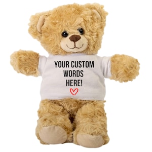 May include: A plush teddy bear with light brown fur and a white t-shirt. The t-shirt has the text "YOUR CUSTOM WORDS HERE!" in black, with a red heart below. The bear has black eyes and a brown nose.