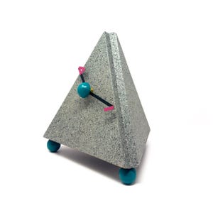 May include: A pyramid-shaped clock with a speckled gray textured finish. The clock face features two thin black hands with small pink rectangular accents at the tips and a central teal sphere. It stands on three round teal ball feet.