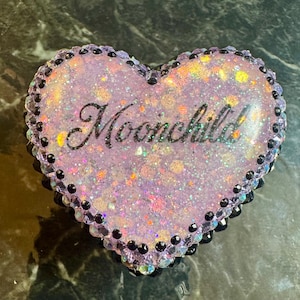 May include: Heart-shaped trinket box with a glittery, light purple surface and the word "Moonchild" in black script. The edges are lined with black and clear beads. The box is a decorative item.