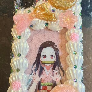 May include: A white and pink phone case with a decorative frame and a picture of an anime character with long black hair. The frame is decorated with flowers, moons, and glitter.