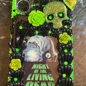 May include: A black and green phone case with a skull, flowers, and a coffin. The case has a picture of a zombie with the text "Night of the Living Dead".