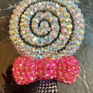 May include: A bedazzled lollipop-shaped accessory. The top is a spiral of iridescent rhinestones with black beads. A pink rhinestone bow is below the lollipop, and the stick is black rhinestones. The accessory is on a dark surface.