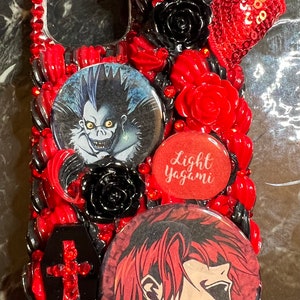 May include: A red, black, and white phone case with a red bow, black roses, and two round buttons featuring characters from the anime Death Note. One button features Light Yagami and the other features Ryuk. The text "Light Yagami" is written on one of the buttons.