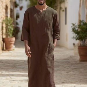 May include: A brown linen robe with a round neckline and three-quarter sleeves. The robe has pockets and is ankle-length. The person is wearing black sandals. The setting is an outdoor walkway.