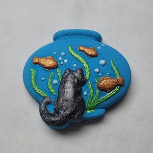 May include: A blue ceramic bowl with a black cat figurine looking at gold fish inside. The bowl has green seaweed and white bubbles painted on it.