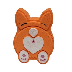 Peut inclure: Bombe de bain orange en forme de fesses de chien de dessin animé avec un cœur blanc et une étoile rose dessus. Le chien a des oreilles roses et des empreintes de pattes roses.