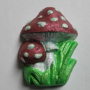 May include: A red and white mushroom with white spots, sitting in green grass. The mushroom is made of a soft, spongy material and has a shiny, iridescent finish.