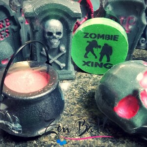 May include: A collection of Halloween-themed bath bombs, including a cauldron, a skull, and a tombstone with the words "Here Lies" on it. The bath bombs are green, black, and red.