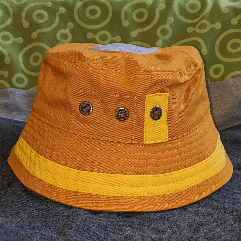 May include: A brown and yellow bucket hat with a gray patch on the top. The hat has three round, black buttons on the front.