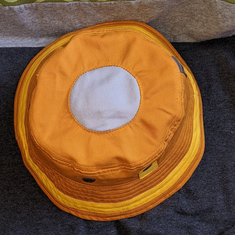 May include: A yellow and orange bucket hat with a light gray circle in the center. The hat has a yellow band around the brim.