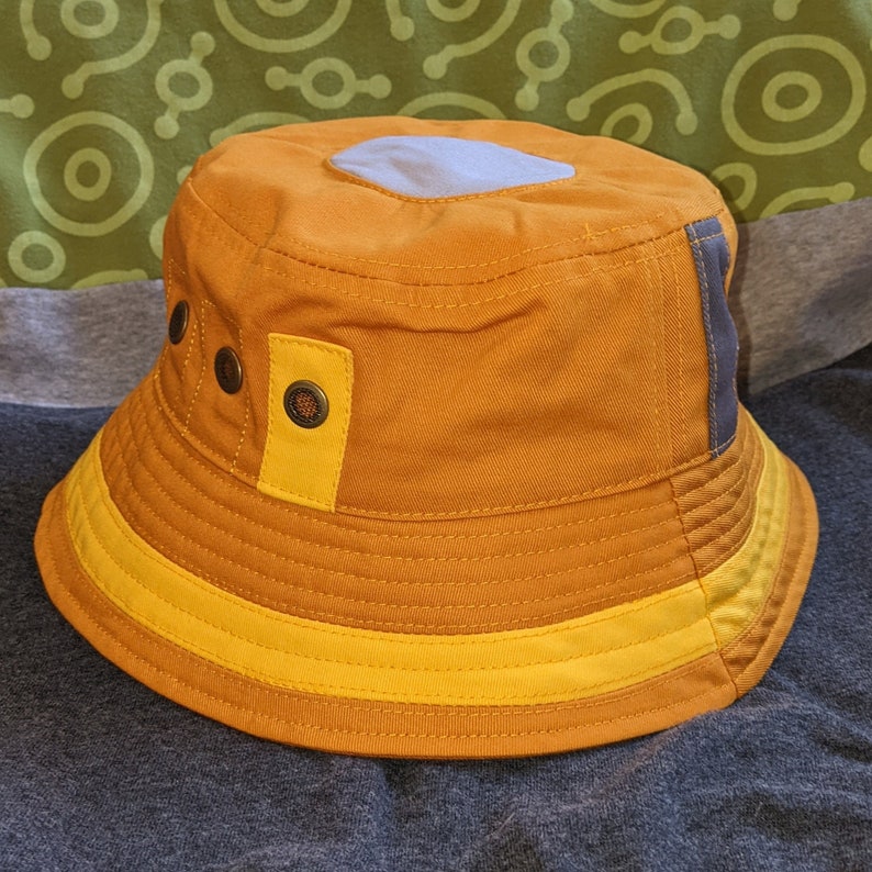May include: A brown and yellow bucket hat with a blue patch on the top. The hat has a yellow band around the brim.