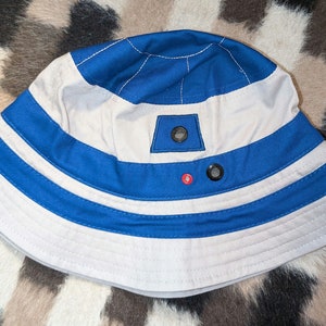 May include: A white and blue bucket hat with a blue and white striped band. The hat has a small, white patch with a black button on the top. The hat is on a brown and white patterned surface.