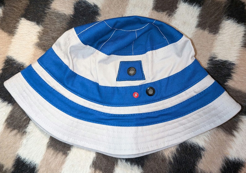 May include: A white and blue bucket hat with a blue and white striped band. The hat has a small, white patch with a black button on the top. The hat is on a brown and white patterned surface.