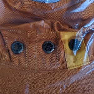 May include: Close-up of a brown canvas hat with a yellow fabric panel and three metal eyelets. The hat has a textured surface and is made of a durable material.