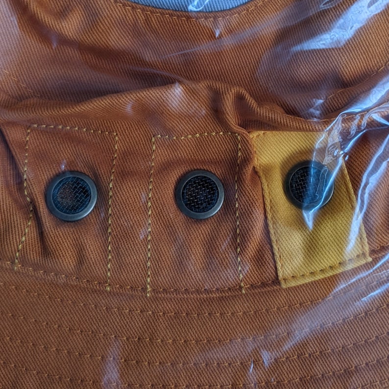 May include: Close-up of a brown canvas hat with a yellow fabric panel and three metal eyelets. The hat has a textured surface and is made of a durable material.