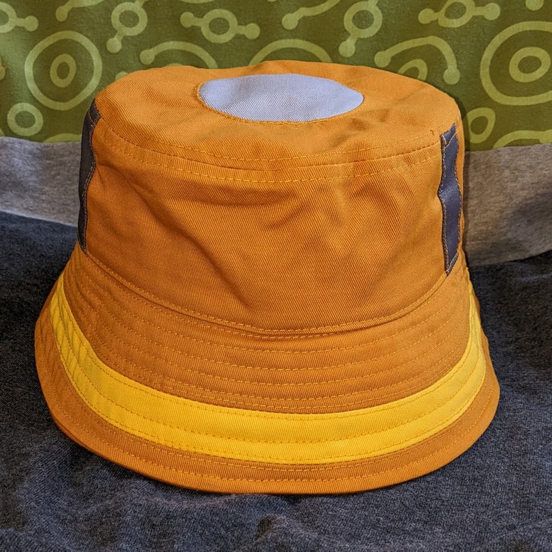 May include: A brown bucket hat with a yellow band and a gray patch on the top. The hat has a dark brown trim around the edge.