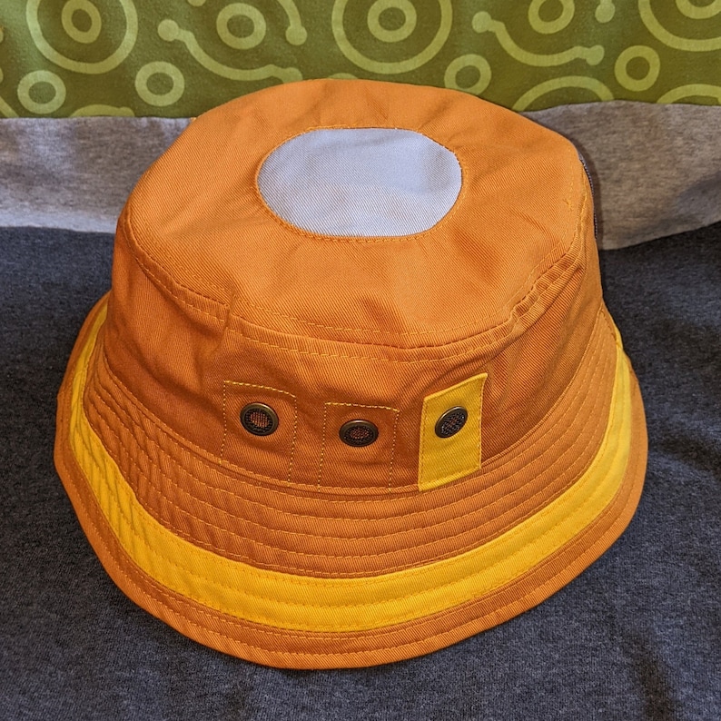 May include: A brown bucket hat with a yellow band and a gray circle on the top. The hat has three metal buttons on the front.