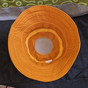 May include: A yellow bucket hat with a brown inner lining and a gray fabric patch on the inside. The hat has a stitched seam around the brim.