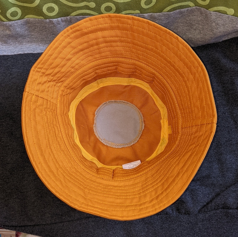May include: A yellow bucket hat with a brown inner lining and a gray fabric patch on the inside. The hat has a stitched seam around the brim.
