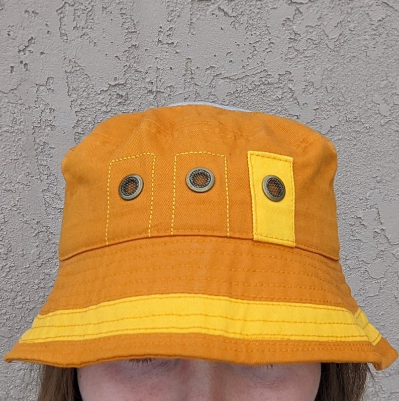 May include: A bright orange bucket hat with a yellow band and three small, round, metal accents. The hat has a wide brim and is made of a soft, textured fabric.
