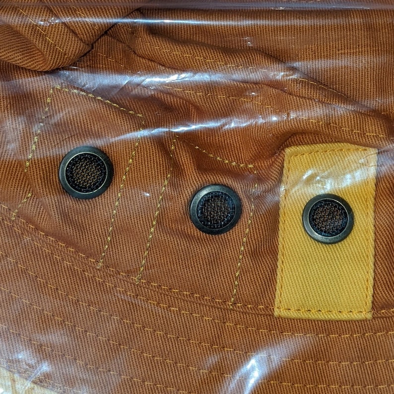 May include: Close-up of a brown and yellow fabric hat with three black metal eyelets. The hat has a textured surface and is made of a durable material.