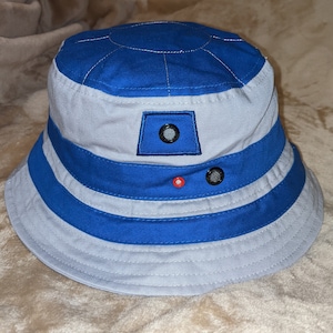 May include: A blue and gray bucket hat with a white and blue striped band. The hat has a small blue patch with a white circle in the center.
