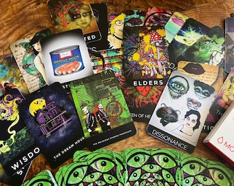 Puede incluir: Una colección de cartas de tarot coloridas con varias ilustraciones, que incluyen una casa, hongos y una calavera. Las cartas están dispuestas sobre una superficie de madera. El texto en las cartas incluye "Wisdom", "Elders", "The Dream House", "The Religious Studies Professor", "Dissonance" y "Oracle Meats".