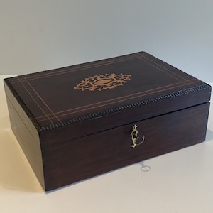 Può includere: Una scatola di legno marrone scuro con un disegno intarsiato sul coperchio. La scatola rettangolare ha una chiusura e una serratura in ottone. Il coperchio presenta un motivo floreale centrale e dettagli del bordo. La scatola è lunga circa 25 cm.