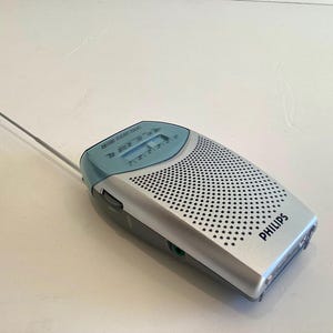 May include: A silver and blue Philips AM/FM radio with a retractable antenna. The radio has a perforated speaker cover and a digital display. The Philips logo is visible on the front.
