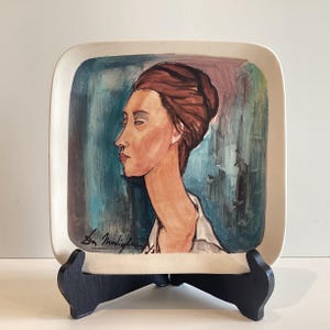 Puede incluir: Plato cuadrado de cerámica con un retrato de una mujer de perfil, con un moño castaño, pintado en tonos marrones, rosas y azules. El plato se exhibe sobre un soporte de madera negro. La firma del artista es visible.