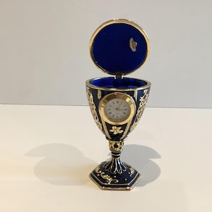 May include: A blue and gold decorative egg-shaped music box with a clock. The egg has a hinged lid lined with blue velvet. The clock face is white with gold hands and Roman numerals. The base is a goblet shape.