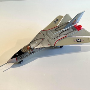 Puede incluir: Un modelo de avión plateado y gris con el texto U.S. AIR FORCE en el lateral, USAF en el ala y marcas F-111A. La aeronave tiene una punta negra, una cabina transparente, escapes de motor rojos y un emblema de estrella.