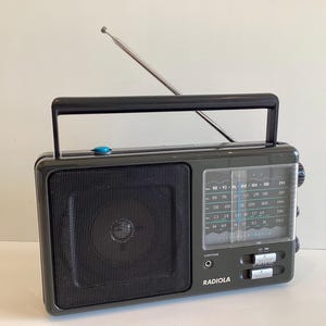 Könnte beinhalten: Ein Vintage-Radiola-Radio in Dunkelgrau mit schwarzem Griff und Antenne. Das Radio verfügt über einen Lautsprecher, ein Einstellrad und Bedienknöpfe. Das Wort "RADIOLA" ist auf der Vorderseite aufgedruckt.