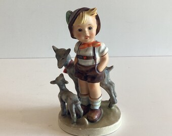Puede incluir: Figura de cerámica de un niño con vestimenta tradicional, de pie con dos pequeñas cabras. El niño lleva un sombrero marrón, pantalones cortos y tirantes, con una pajarita roja. Las cabras son grises y están sobre una base circular.