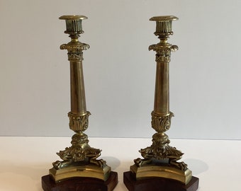 May include: Pair of antique brass candlesticks with ornate detailing. Each candlestick features a fluted column, decorative base, and a wooden base. The brass has a warm, golden hue, and the wooden base is a dark brown color.