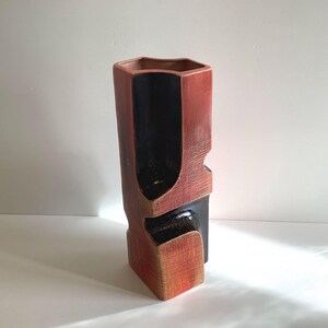May include: A tall, geometric ceramic vase featuring a textured, reddish-orange exterior finish and a contrasting smooth, deep black interior. The structure has an angular, multi-faceted form with recessed sections and a hexagonal top opening.