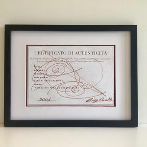 May include: Framed certificate of authenticity with a black frame and white matting. The certificate has text in Italian and a signature. The frame is rectangular and sits on a white surface.
