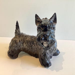 Puede incluir: Figura de perro Scottish Terrier de cerámica negra brillante. El perro está representado sentado, con la cola levantada y las orejas erguidas. La figura tiene una textura de pelaje detallada.