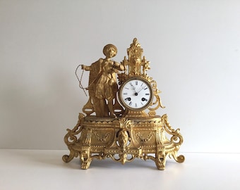 May include: A gold-toned mantel clock featuring a central white circular dial with black Roman numerals and hands. The clock is adorned with an ornate metallic figure of a person holding a rope, set upon a decorative rectangular base with intricate relief carvings and scrollwork details.