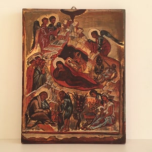 May include: A vintage religious icon depicting the Nativity scene. The artwork features figures in red, brown, and gold tones, with angels and other biblical figures. The icon is mounted on a wooden panel with a gold border.