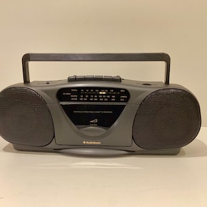 May include: A gray RadioShack boombox with a handle, two speakers, and a radio dial. The boombox has a cassette recorder and the RadioShack logo. The speakers are dark gray and the body is a lighter gray.