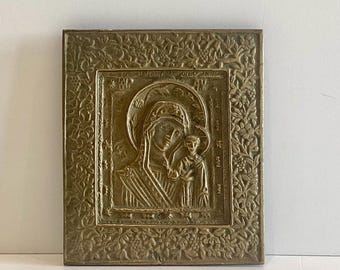 Puede incluir: Icono religioso antiguo de latón, de forma rectangular, que representa a la Virgen María y el niño en relieve. La imagen central está enmarcada por un borde floral, con detalles intrincados y un aspecto vintage.