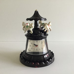 Könnte beinhalten: Eine schwarze Karusselluhr mit Quarzwerk. Die Uhr hat eine rotierende Oberseite mit drei weißen Pferden mit goldenen Akzenten. Das Zifferblatt ist weiß mit schwarzen Zahlen und Zeigern. Ein dekorativer Artikel.