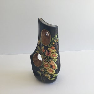 May include: A black decorative vase with a unique shape, featuring two circular cutouts and a floral design. The vase has a glossy finish and a wooden accent. The floral design includes yellow and orange flowers with green leaves.